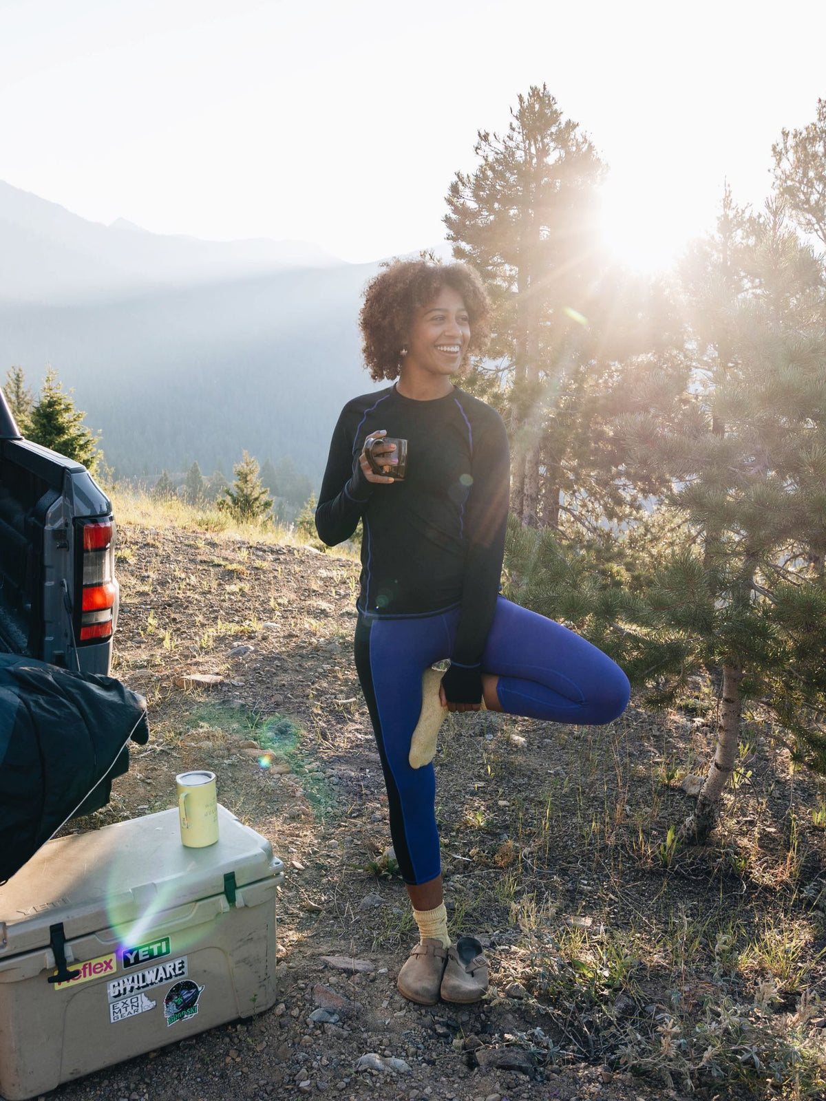 [Black] Woman camping in the mountains at sunrise, wearing a black Bassett LITE Raglan and holding a mug