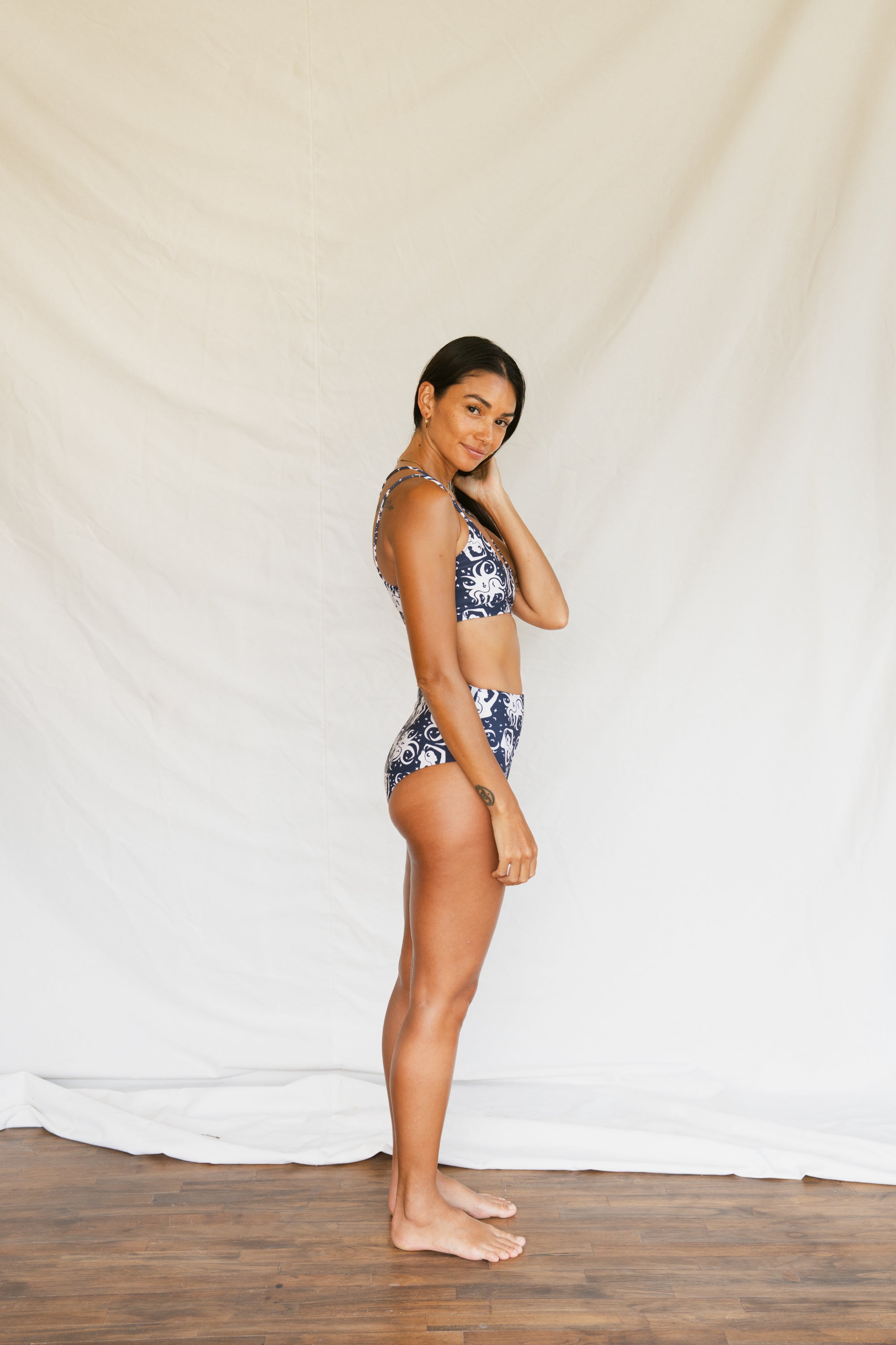 Person wearing a navy blue bikini with white octopus-like pattern on a plain background, side full body view