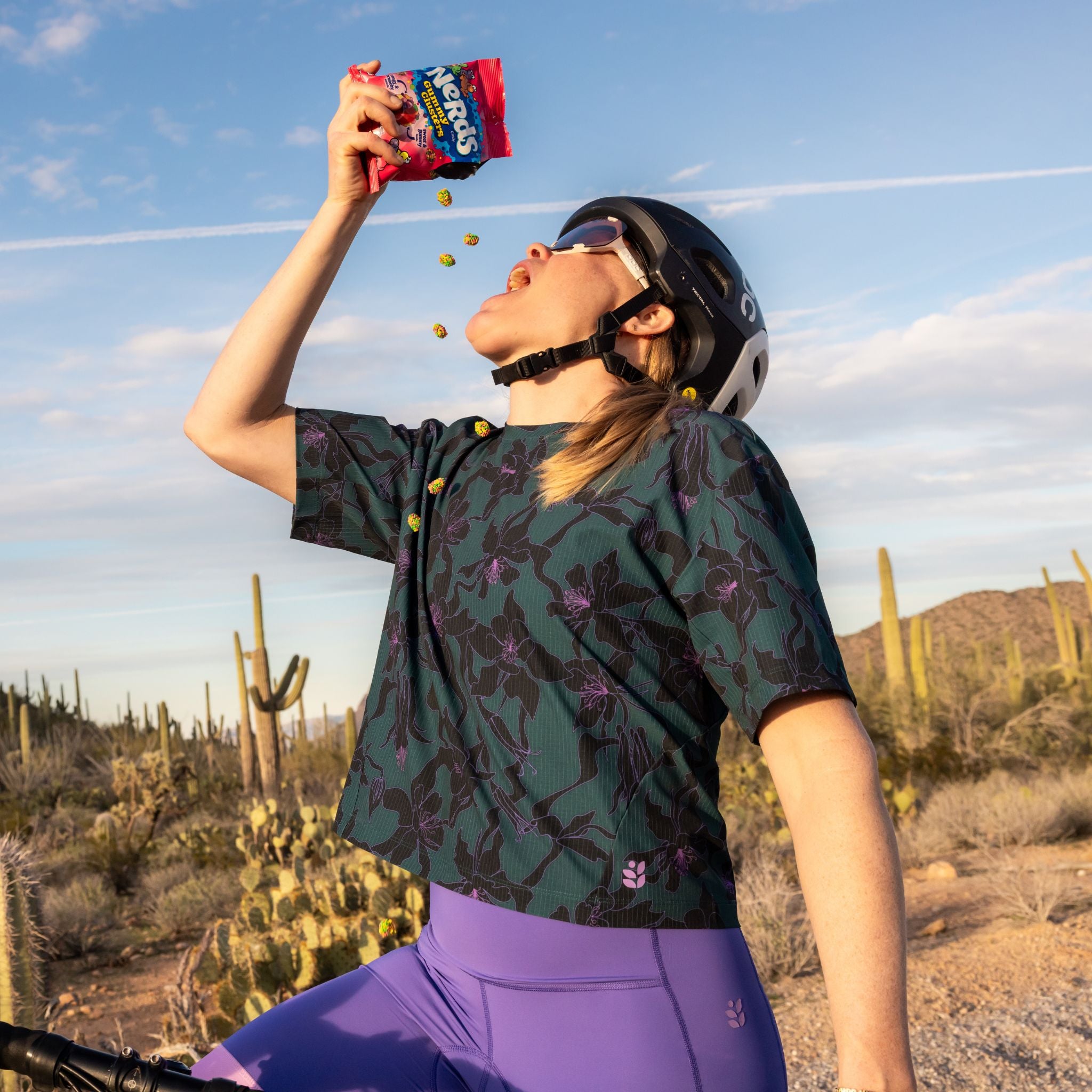 [Spruce Aquila Bloom] Person in a desert setting eating nerds clusters, with cacti and mountains in the background.