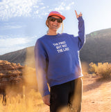 Woman standing in the desert wearing blue sweatshirt with text "the first but not the last"