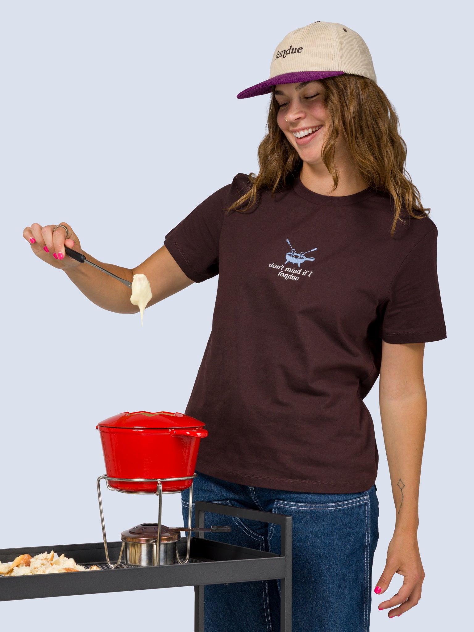 [Fondue] Lifestyle shot of a model wearing the Fondue Graphic Tee in dark brown, dipping bread into a red fondue pot