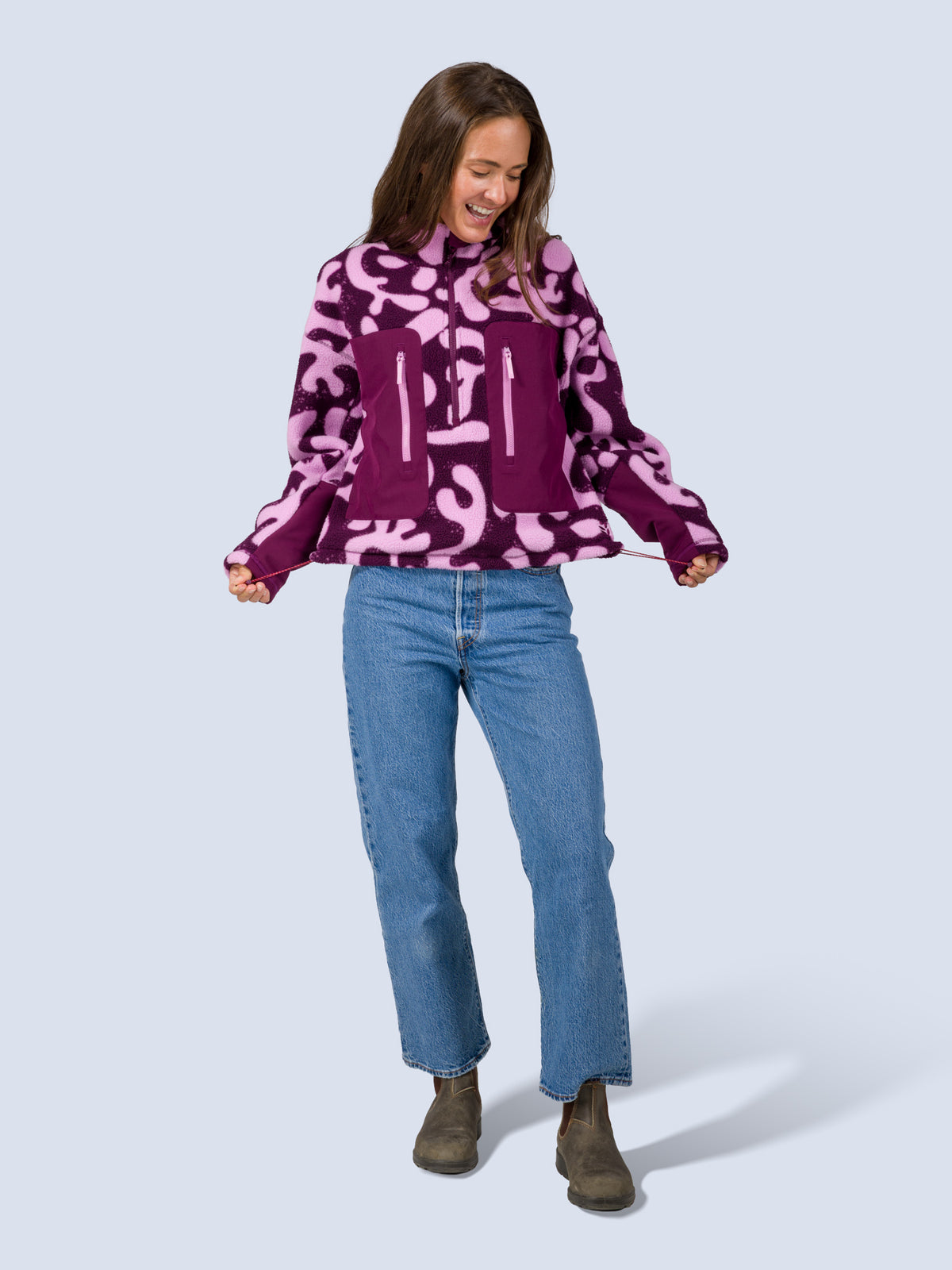 [Mulberry Reef] Model wearing the Banner 3/4 Zip Jacket in purple and pink abstract-print fleece with jeans, laughing while adjusting the hem