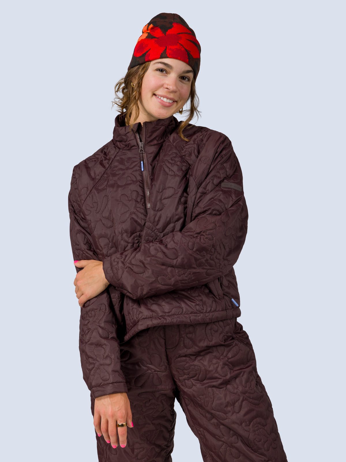 [Dapple] A woman modeling a matching quilted winter jacket and pants set in a deep plum-brown color, paired with a red and black patterned beanie, against a solid light blue-gray background.