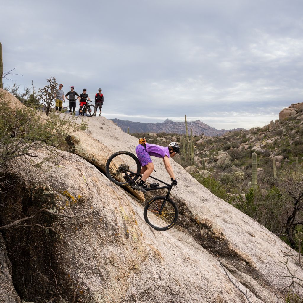 [Amethyst Aquila Bloom] Woman wearing purple bike outfit, riding down rocky trail