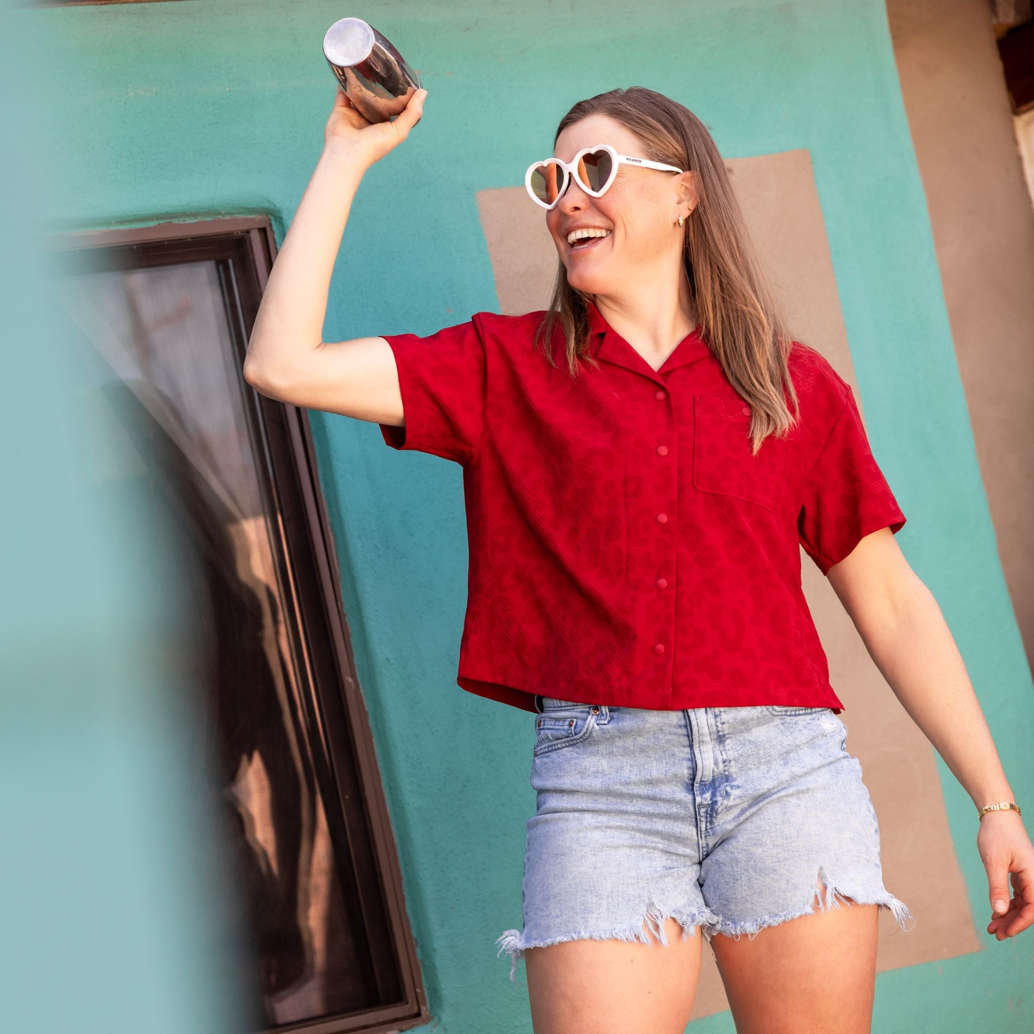 [Garnet Meow] Woman in a red shirt and denim shorts mixing a drink against a teal wall.