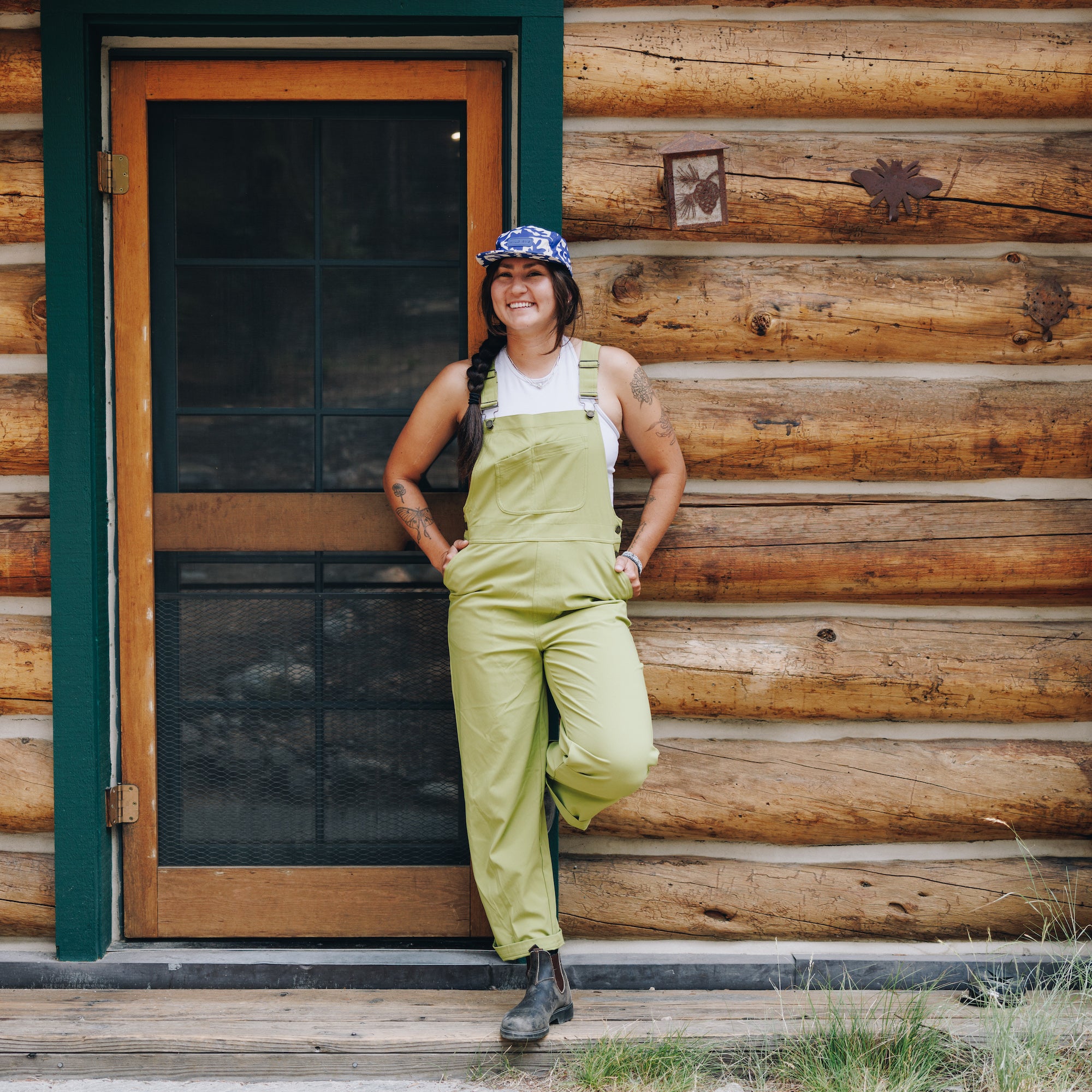 [Caper] Woman leaning against log cabin in green overalls