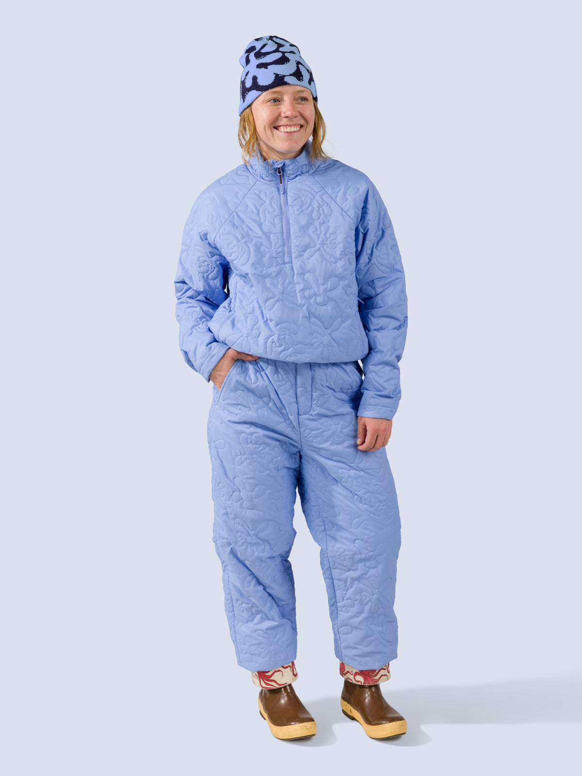 [Blue Reef] A woman wearing a light blue quilted jacket and pants with a matching tamarack toque beanie and brown boots, standing against a light gray studio background.