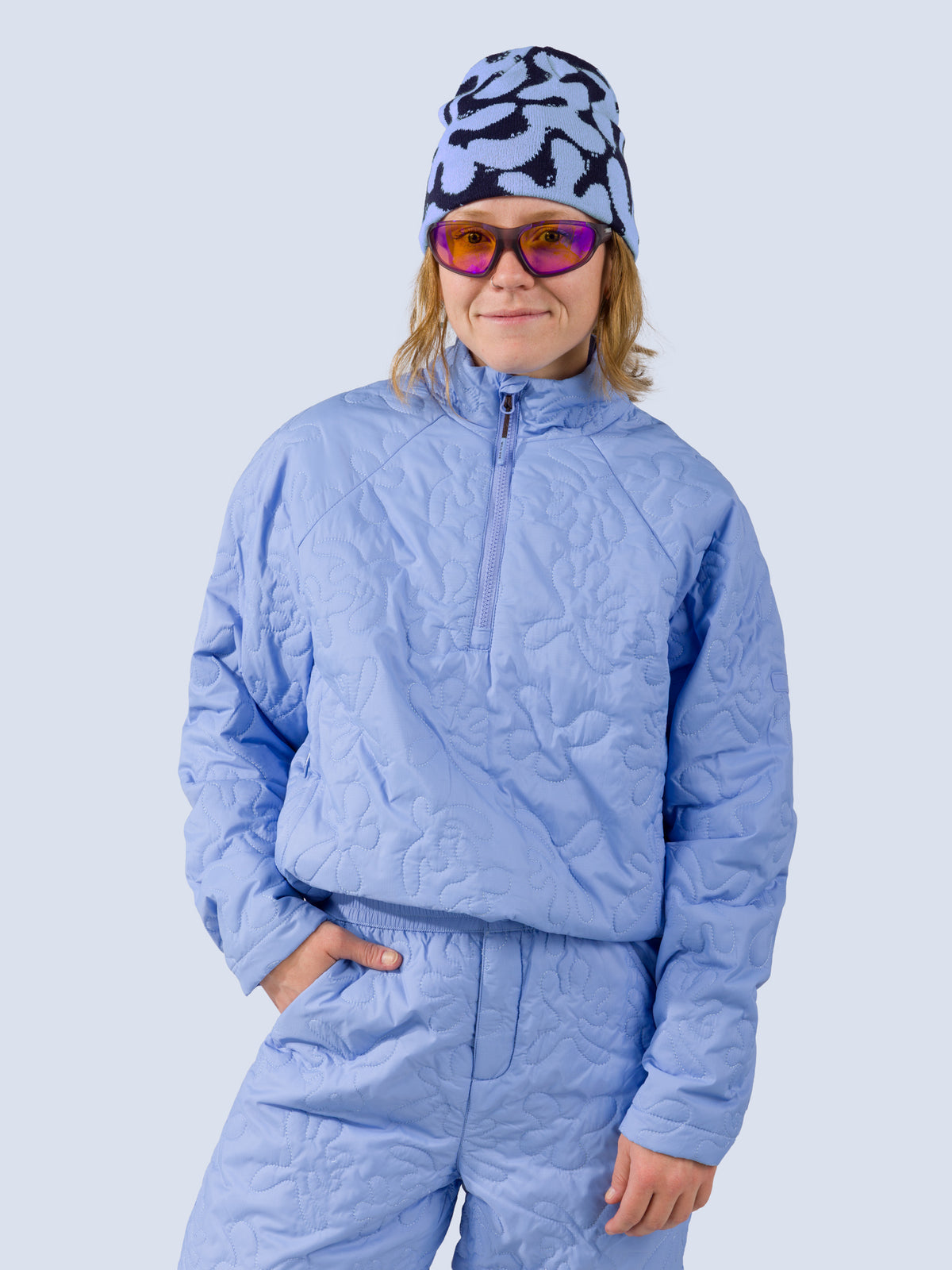 [Blue Reef] A model wearing a light blue quilted half-zip jacket and matching light blue pants, paired with a blue and black patterned beanie and purple-tinted sunglasses, against a solid light gray background.