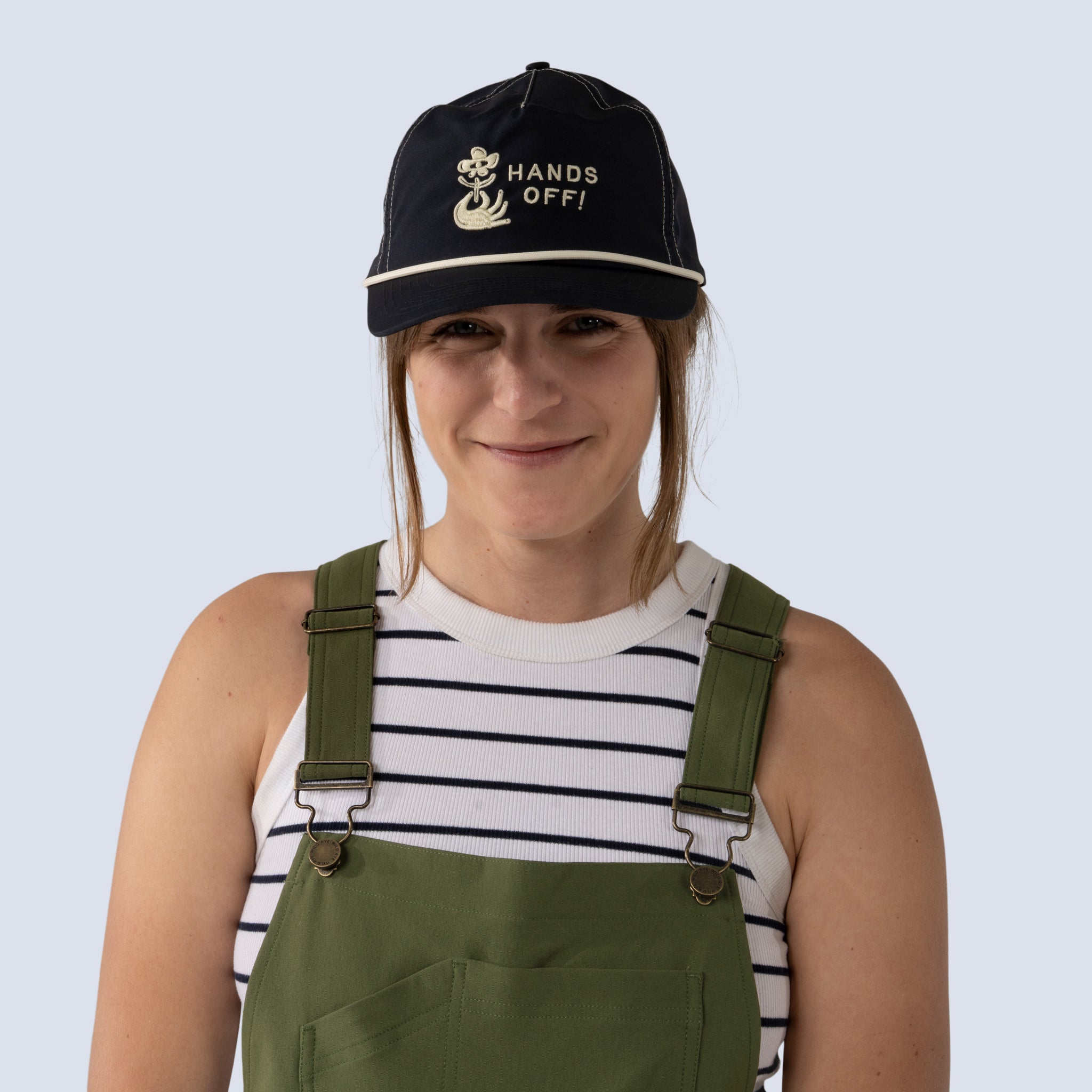 Person wearing a navy cap with 'HANDS OFF!' text, striped shirt, and green overalls 