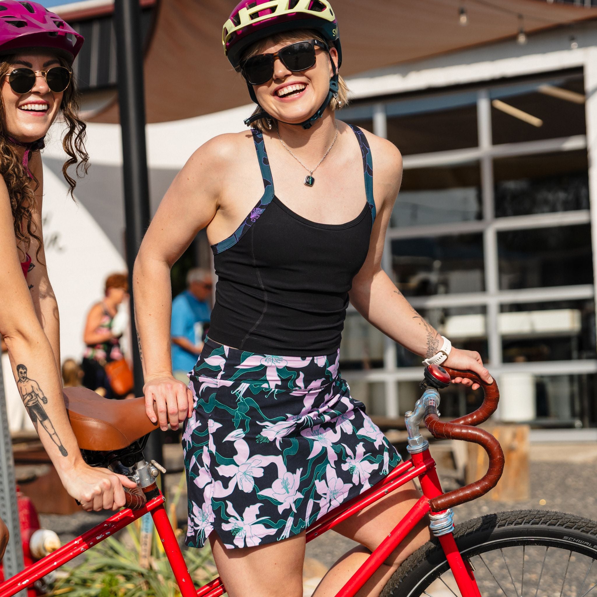 [Black Aquila Bloom]Two women on a tandem bike wearing helmets and sunglasses, with a building in the background.